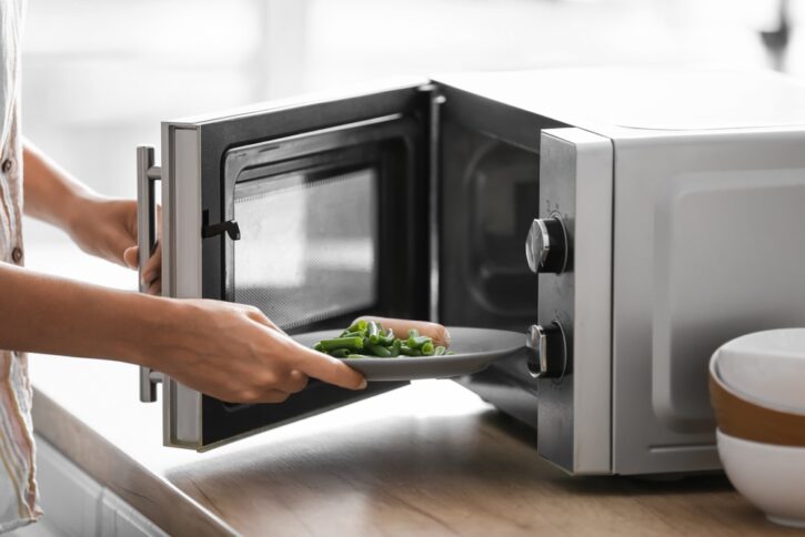 Young,Woman,Putting,Plate,With,Food,Into,Microwave,Oven,In